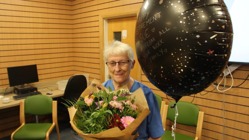 Sue Pounder holding flowers. She stands next to a balloon which reads: "Happy retirement Sue, love from all your work family".