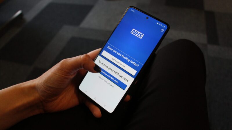 A hand holding a mobile phone. The NHS app is open. It reads: "How are you feeling today? Get health advice." Below reads: "To access your NHS Services. Continue with NHS login".