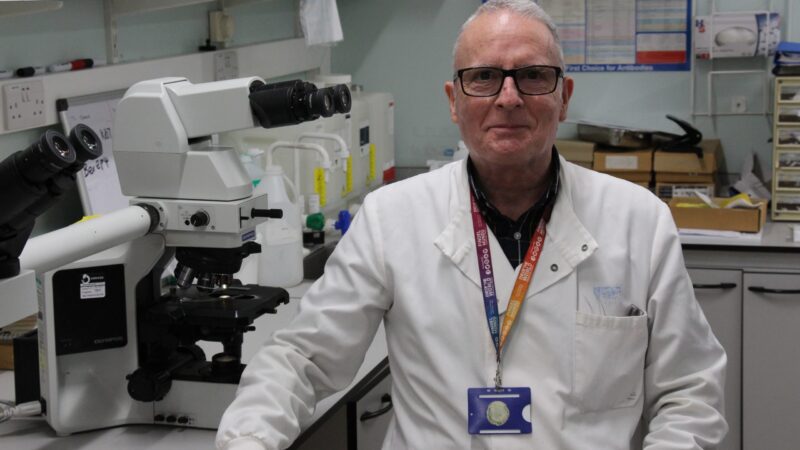 James France in a pathology lab. He wears a white lab coat and sits with a microscope.