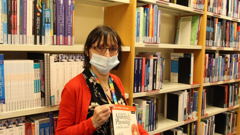 Debbie Elliot holds a textbook. She stands in a library.