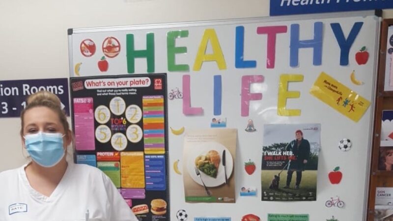 A member of staff in front of a display board. It features posters and information about healthy eating and exercise.