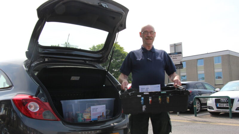 A volunteer driver stands in front of a car. He holds a crate of food.